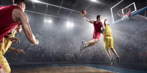  Basketball player makes slam dunk on big professional arena. Player flies through the air with the ball. Opponents try to prevent the ball from hitting the basketball ring. © Alex