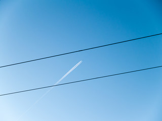 In the clear blue sky, a high flying airplane leaves condensation trail behind itself.	 The sky is crossed by a line of wires.