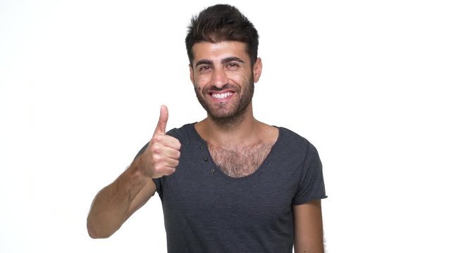 hispanic young man wearing grey t-shirt looking at camera smiling broadly with white teeth showing thumb up over white background. Concept of emotions