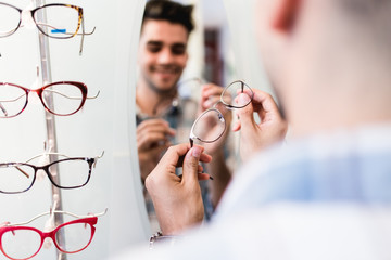Handsome young man choosing eyeglasses frame in optical store.