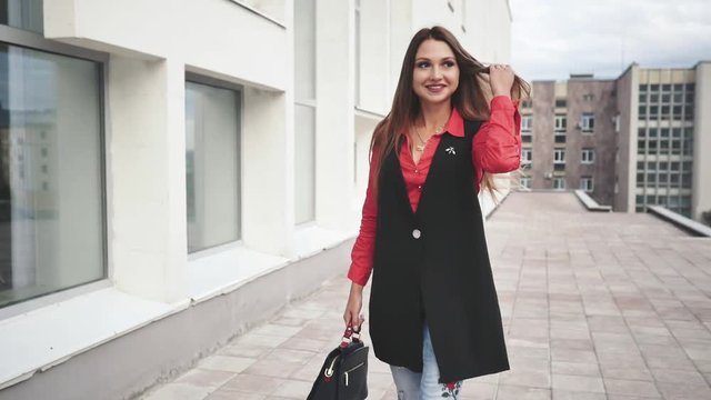 the happy young woman with long hair in a red shirt and a long sleeveless jacket walks down the street