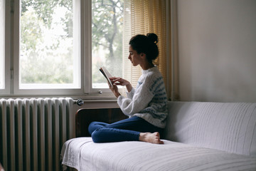 Young beautiful woman reading a book on a couch near a window