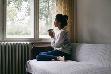 Young beautiful woman holding a cup of coffee on a couch near a window