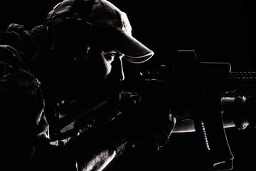 Studio contour backlight shot of special forces soldier in uniforms and baseball cap, pointing rifle, closeup portrait on black background