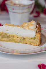 A piece of pie with rhubarb and meringue  is on a white table near red roses and cups of tea up close.