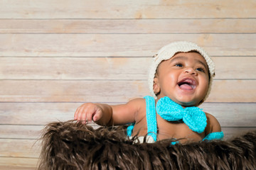 Infant mixed race boy wearing hat and bow tie sitting in a fluffy furry basket wooden background...