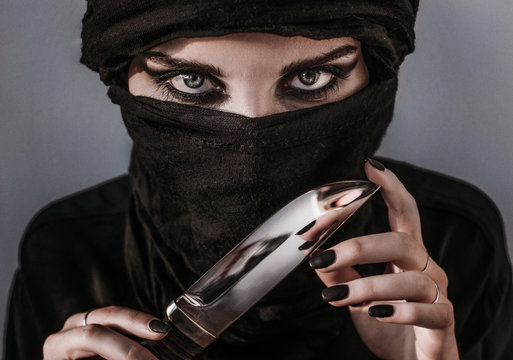 Woman wearing hijab with smokey eyes make up looks straight in camera holding knife in front of her face. Threat with strong look.