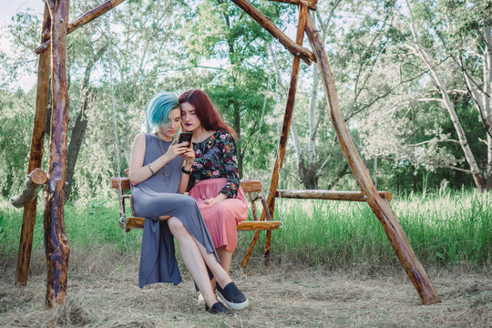 Two girls stits on wooden swing, looking at smartphone. Browsing internet, posting photos in social media