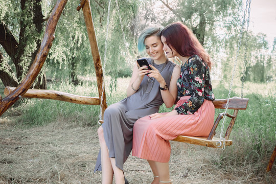 Two girls stits on wooden swing, looking at smartphone. Browsing internet, posting photos in social media