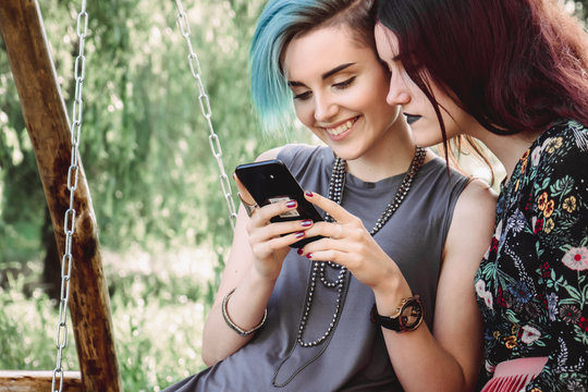 Two girls stits on wooden swing, looking at smartphone. Browsing internet, posting photos in social media