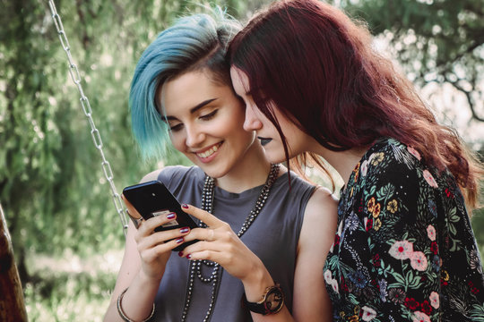 Two girls stits on wooden swing, looking at smartphone. Browsing internet, posting photos in social media