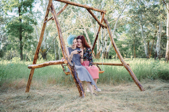 Two girls stits on wooden swing, looking at smartphone. Browsing internet, posting photos in social media