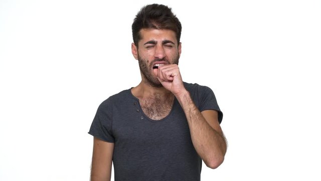 picture of handsome young man in grey t-shirt looking at camera yawning covering mouth with fist being sleepy over white background. Concept of emotions