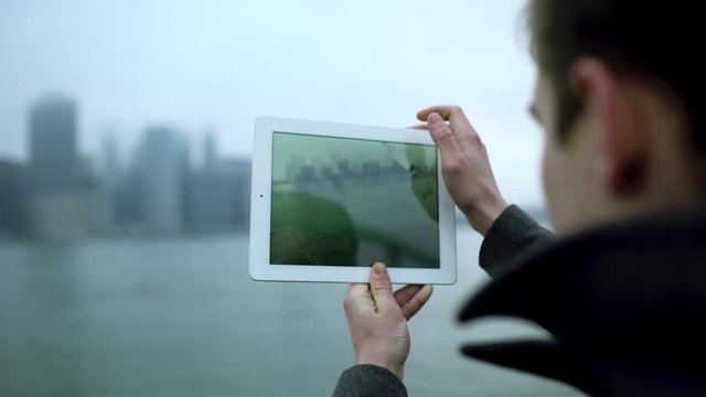 Young tourist taking a photo of Manhattan island on his tablet pc while standing near Hudson river