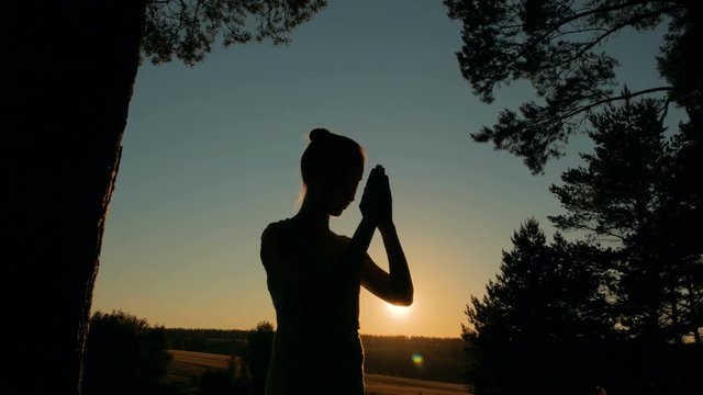 Silhouette of young woman praying in forest at sunset. Sunset light, sun lens flares, golden hour. Religion, spiritual and nature concept