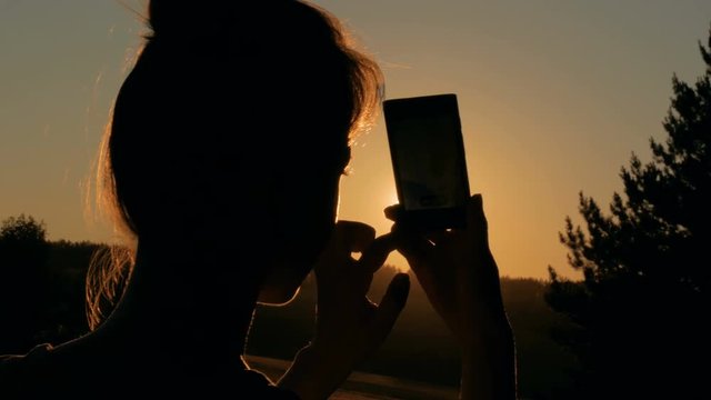 Woman silhouette taking photo of beautiful sunset with smartphone in forest. Sunset light, golden hour, lens flares. Close up shot of woman hands with mobile. Photography, nature and social concept