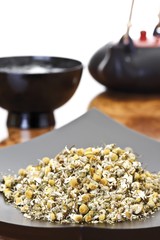 Dried chamomile blossoms in a black bowl, tea served in an Asian tea set