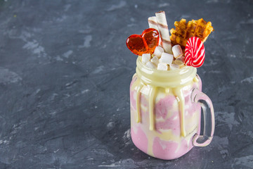 Freakshake from pink smoothie, cream. Monstershake with lollipops, waffles and marshmelow. Extreme milkshake in a Mason jar. Gray dark concrete background.