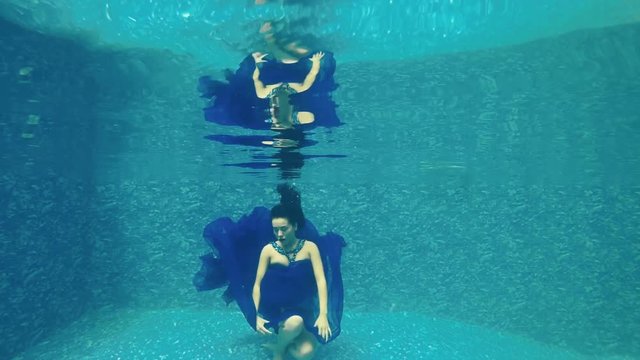 Beautiful girl in long blue dress poses for camera underwater in pool