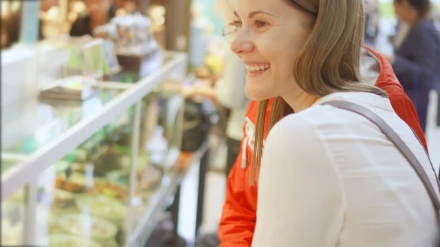 Senior woman in red coat and her daughter in white shirt shopping in mall. Standing looking at glass case discussing purchases