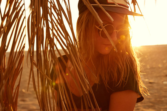 Portrait of a girl in sunset in contra light in palm leaves on the beach at the sea in sunglasses and baseball cap