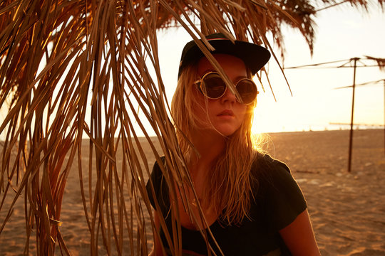 Portrait of a girl in sunset in contra light in palm leaves on the beach at the sea in sunglasses and baseball cap