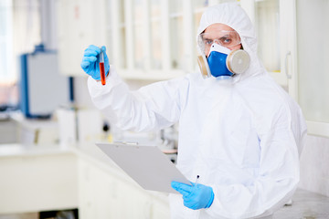 Waist-up portrait of confident virology research scientist posing for photography with test tube in hand while developing vaccine and taking notes on clipboard, interior of modern office on background