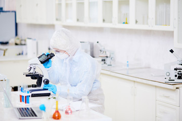 Profile view of female microbiologist wearing hazmat suit looking through microscope while studying...