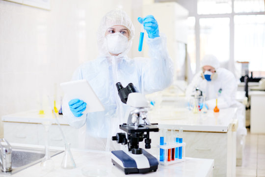 Gifted young microbiologist wearing filtering mask and coverall using digital tablet while carrying out experiment with biohazard substance, interior of spacious laboratory on background