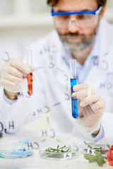 Concentrated bearded microbiologist wearing white coat and safety goggles holding test tubes with...