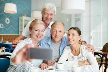 Two smiling senior couples taking selfie on smartphone while gathered together at lovely small coffeehouse