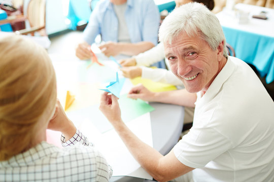 Portrait Shot Of Cheerful Senior Man Looking At Camera With Toothy Smile While Making Origami Figure From Colorful Paper, His Friends Sitting Next To Him