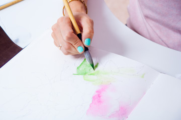 Close-up of a female artist paints a thin wooden brush and watercolor drawing of a pink peony flower in an album for drawing on a white background