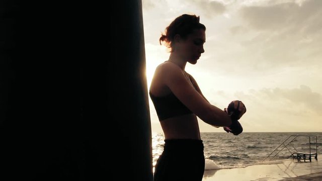 Camera round movement: female boxer wrapping hands with boxing wraps on the beach by the ocean standing against the sun. Lens flare