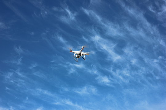 White quadrocopter is flying high in the air, taking photos and recording footage from above. Flying drone with four motors and propellers, camera and red warning lights on clear blue sky background.