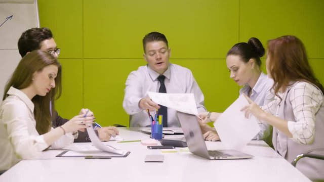 Business teamwork of young managers and chief during serious and active scrum meeting, working with papers and reports, sitting in modern green office room