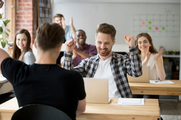 Young casually dressed male office worker throws hands in air celebrating achievement at work. Coworkers around cheer and clap hands. Rewarding outcome, received promotion, achieved success concept.