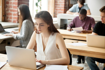 Casual business woman typing on laptop in modern shared office surrounded by coworkers. Young female entrepreneur composing email, communicates with clients or business partners, setting appointments.
