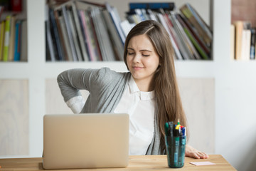 Young tired female office worker having back discomfort at work desk in office. Casual fatigued businesswoman feeling back pain after sitting on uncomfortable chair in front of laptop for long hours.