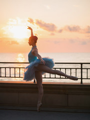 Dancing ballerina in blue ballet tutu and point on embankment above ocean or sea at sunset. Young...