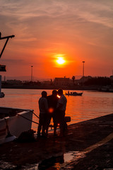 Three fishermen on the pier in the rays of the setting summer sun. Sunset at the port for yachts in Italy, Ortigia, Siracusa