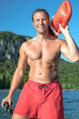 young, sexy, muscular man is doing lifeguard - he is watching at the bay for the safety. he wears red swimming trunks and is holding an red rescue buoy and a binoculars. 