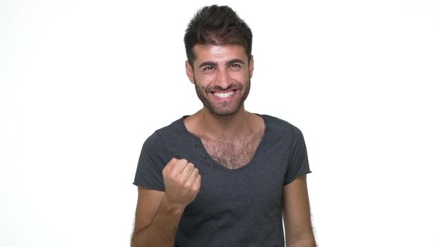 picture of hispanic young man wearing grey t-shirt looking at camera enjoying success with clenched fist over white background slow motion. Concept of emotions