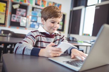 Smiling schoolboy using laptop and mobile phone on desk at