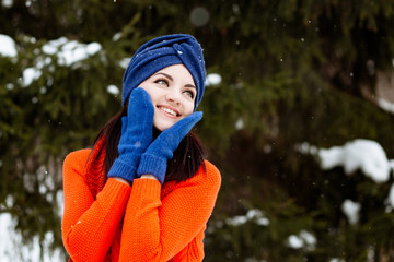 Outdoor winter portrait of beautiful happy smiling young woman posing on street. The girl is dressed in a bright orange sweater and a blue cap
