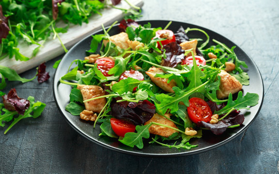 Fresh salad with chicken breast, arugula, nuts and tomatoes on black plate in a wooden table.