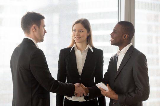 African american and caucasian business partners shaking hands after successful negotiation in office. Young female secretary standing between two company leaders welcoming each other before meeting