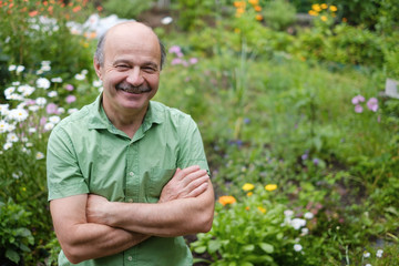 Naklejka na ściany i meble An elderly man with a mustache and a bald spot in a green T-shirt is standing among flowers in the summer garden, arms crossed and smiling.