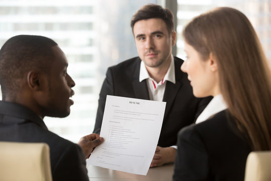 Rear view african american company leader analyzing and discussing applicants resume with female hr specialist, caucasian male job candidate waiting for hiring decision after interview on background