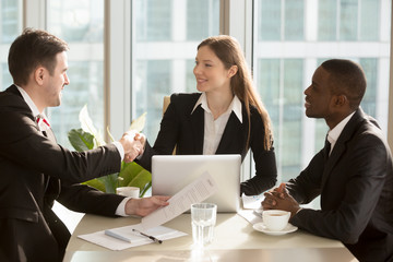 Happy female company leader sitting at desk with her african american partner, shaking hand with satisfied businessman after successful negotiations and contract conclusion. International cooperation